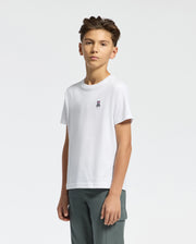 A young person with short brown hair wears the KIDS CLASSIC CREW NECK TEE by Psycho Bunny, showcasing the embroidered Classic Psycho Bunny logo, paired with dark green pants against a plain light background.