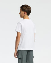 A young person with short brown hair stands sideways wearing Psycho Bunny's Kids Classic Crew Neck Tee in 100% Pima cotton and gray cargo pants. The plain white shirt showcases the classic embroidered Psycho Bunny logo on a neutral background.