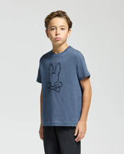 A young boy with light brown hair stands against a plain background, wearing a Psycho Bunny KIDS MONARC FRONT BUNNY GRAPHIC TEE - B0U211E200, made from soft Pima cotton. The blue t-shirt showcases a raised-printed bunny face over crossed bones. He pairs it with black pants and gazes directly at the camera.