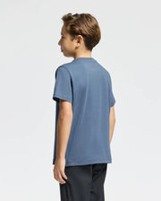 A young boy with light brown hair stands facing away, wearing a dark blue Kids Monarc Front Bunny Graphic Tee by Psycho Bunny, crafted from soft Pima cotton, coupled with black pants. The raised-printed Bunny design introduces a playful element against the plain white background.