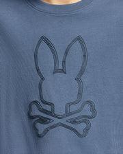 A close-up of the Kids Monarc Front Bunny Graphic Tee by Psycho Bunny, product code B0U211E200, in blue reveals a stylized, raised-printed bunny head above crossed bones, emulating a skull and crossbones motif. The design is crafted on premium Pima cotton in a slightly darker shade for subtle contrast.