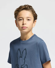 A young boy with light brown hair stands against a plain background, wearing a KIDS MONARC FRONT BUNNY GRAPHIC TEE by Psycho Bunny. The blue T-shirt, crafted from soft Pima cotton, showcases a prominent raised-printed bunny design in the center. He gazes directly at the camera with a neutral expression.