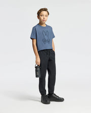 A young boy stands against a simple background wearing a blue KIDS MONARC FRONT BUNNY GRAPHIC TEE - B0U211E200 by Psycho Bunny, crafted from soft Pima cotton. He pairs it with black joggers and sneakers while holding a black water bottle in his left hand.