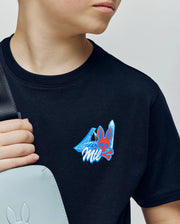 A child wears the Psycho Bunny KIDS MONTREAL TEE (B0U381F200), a black 100% Pima cotton shirt with a blue and red bunny skull, crystals, and "Mil" graphic, paired with a light gray bag featuring a subtle bunny logo.