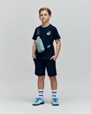 A young boy stands against a plain background wearing the Psycho Bunny KIDS MONTREAL TEE (B0U381F200), black shorts, blue sneakers, striped socks, and a gray crossbody bag. He looks straight ahead with hands in his pockets.
