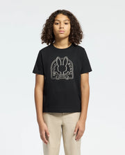 A person with curly hair stands against a plain background, wearing the KIDS MELBOURNE GRAPHIC TEE by Psycho Bunny, featuring a bunny graphic and "Bone Bunny House" in white. They pair it with eco-friendly beige pants.