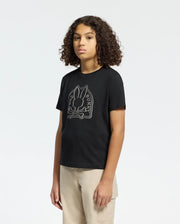 Someone with curly hair is wearing a Psycho Bunny Melbourne graphic tee made from eco-friendly Peruvian Pima cotton. The black tee has a bunny with a bow tie design and "Bunny" text. They wear it with light-colored pants against a plain, light background.