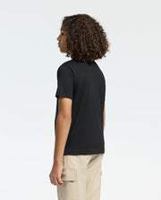 Someone with curly hair is wearing a Psycho Bunny KIDS MELBOURNE GRAPHIC TEE (B0U410F200) in black and beige cargo pants, standing against a plain white backdrop, showcasing an eco-friendly style.