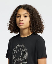 A person with curly hair wears a black KIDS MELBOURNE GRAPHIC TEE (B0U410F200) by Psycho Bunny, crafted from eco-friendly Peruvian Pima cotton and featuring a bunny logo with bones. They look ahead neutrally against a plain backdrop.