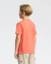 A young blonde-haired boy faces away, wearing a Psycho Bunny KIDS MELBOURNE GRAPHIC TEE in bright orange, made from soft Pima cotton and featuring a charming bunny design. He pairs it with beige cargo shorts against a plain white background.