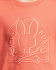 Close-up of a coral-colored KIDS MELBOURNE GRAPHIC TEE by Psycho Bunny, showcasing a raised Bunny graphic with a stylized bunny head above crossed bones and "Psycho Bunny" written along the sides.