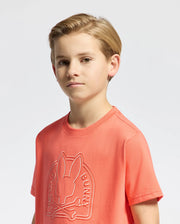 A young boy with short blonde hair sports a Psycho Bunny KIDS MELBOURNE GRAPHIC TEE - B0U410F200 in salmon. Made from soft Pima cotton, it features the iconic stylized bunny and crossbones logo on a plain light gray background.