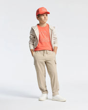 A young boy stands confidently in a beige hoodie and joggers with hands in pockets. He wears the "Kids Melbourne Graphic Tee" by Psycho Bunny, featuring a whimsical bunny graphic, and complements it with white sneakers and a bright coral hat against a plain white background.