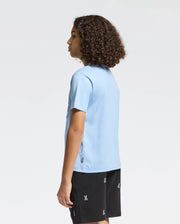 With curly hair and standing in a partial side view against a plain background, the person is wearing an eco-friendly light blue Psycho Bunny KIDS MELBOURNE GRAPHIC TEE - B0U410F200, made of 100% Pima cotton, paired with black shorts.