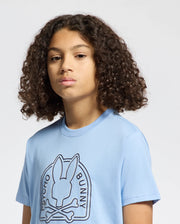 A curly-haired individual is wearing a Psycho Bunny KIDS MELBOURNE GRAPHIC TEE (B0U410F200) in light blue, made from 100% Pima cotton. The tee features an eco-friendly bunny and crossbones design, set against a plain background.