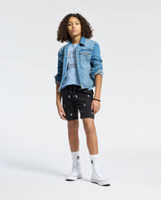 A teenager with curly hair models a Psycho Bunny KIDS MELBOURNE GRAPHIC TEE made from 100% Pima cotton. Standing on one leg in black shorts with bunny prints, they wear white socks and high-top sneakers, hands in pockets against a plain backdrop, highlighting an eco-friendly style.