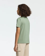 A curly-haired person stands facing away, wearing a Psycho Bunny green Pima cotton T-shirt with a subtle Bunny graphic and beige pants with side pockets. The background is plain light gray. Product: KIDS MELBOURNE GRAPHIC TEE - B0U410F200.