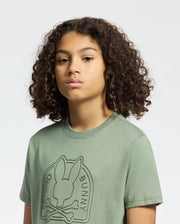 A person with curly hair wears the KIDS MELBOURNE GRAPHIC TEE by Psycho Bunny, featuring a bold bunny graphic with a crossbones logo on soft green Pima cotton. The plain light-colored background highlights the shirt's unique design.