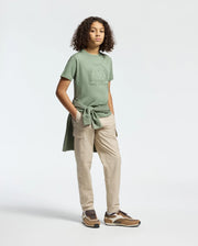 In a simple studio, a curly-haired person wears a casual menKnit outfit with the Psycho Bunny KIDS MELBOURNE GRAPHIC TEE in green, light khaki cargo pants, brown sneakers, and ties a Pima cotton green sweatshirt around their waist.