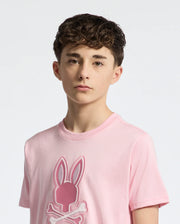 A young person with short, curly brown hair wears a light pink Psycho Bunny KIDS NEW CASTLE EMBROIDERED GRAPHIC TEE featuring a stylized bunny and crossbones logo, made from premium Pima cotton. They have a neutral expression against a plain light background.