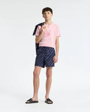 A teen in a pink Psycho Bunny KIDS NEW CASTLE EMBROIDERED GRAPHIC TEE stands holding a jacket over his shoulder. Dressed in blue patterned shorts and black slide sandals, he poses against a plain white background, effortlessly blending comfort with style.