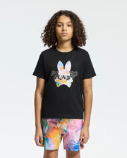 A person with curly hair is wearing a Psycho Bunny KIDS MILOS GRAPHIC TEE - B0U456F200 in black, featuring a vibrant space-age bunny design crafted from luxurious Peruvian Pima cotton, paired with multicolored abstract pattern shorts, standing against a plain background.