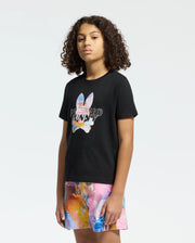 A young person with curly hair wears a black Psycho Bunny KIDS MILOS GRAPHIC TEE, showcasing a vibrant space-age design and the word "PUMA." Paired with multicolored shorts, they stand against a simple white background.