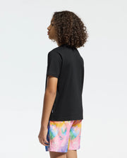 A curly-haired person wears a Psycho Bunny KIDS MILOS GRAPHIC TEE – B0U456F200, a black t-shirt with a Center Bunny Graphic. They pair it with colorful shorts featuring pink, blue, yellow, and orange hues. The vibrant pattern contrasts against the plain background as they pose slightly to the side.