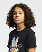 A young person with curly hair wears a black KIDS MILOS GRAPHIC TEE by Psycho Bunny, crafted from soft Peruvian Pima cotton. It features a colorful, space-age bunny graphic and text "Psycho Bunny," all on a plain white background enhancing the vibrant design.