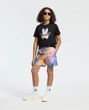 A person with curly hair wearing sunglasses and a Psycho Bunny KIDS MILOS GRAPHIC TEE in black confidently dons vibrant multicolored shorts, white socks, and sleek white sneakers.