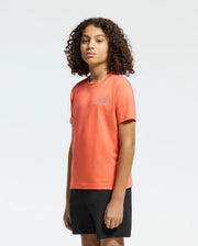 A young person with curly brown hair wears the Psycho Bunny KIDS SYDNEY BACK GRAPHIC TEE - B0U509F200, a regular fit orange Pima cotton shirt, paired with black shorts, standing against a plain light background and facing forward neutrally.