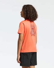 A curly-haired person faces away from the camera in a Psycho Bunny KIDS SYDNEY BACK GRAPHIC TEE (B0U509F200), an orange regular-fit Pima cotton t-shirt with a large bunny skull and crossbones print on the back, paired with black shorts.