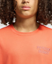 A child with curly hair wears the Psycho Bunny KIDS SYDNEY BACK GRAPHIC TEE in orange, featuring a water-based "PSYCHO BUNNY" print on the left chest. The photo is cropped to show only the lower face and upper torso.