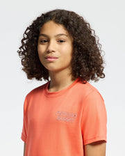 A young person with curly brown hair wears the Psycho Bunny KIDS SYDNEY BACK GRAPHIC TEE (B0U509F200) in coral and looks confidently at the camera, standing against a plain light background.