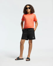 A young person with curly hair, wearing sunglasses, black shorts, and black slide sandals stands confidently with hands in pockets against a plain white background, showcasing the Psycho Bunny KIDS SYDNEY BACK GRAPHIC TEE - B0U509F200.