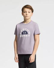 A young person poses against a plain background wearing the KIDS GOLDIE GRAPHIC TEE by Psycho Bunny, featuring the bunny graphic. The light purple tee, crafted from Peruvian Pima cotton, pairs well with their short light brown hair as they gaze directly at the camera.