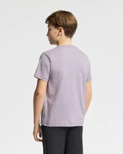 A young boy with short hair stands facing away, glancing to the side while wearing a light purple Psycho Bunny KIDS GOLDIE GRAPHIC TEE made of soft Pima cotton. The contrast with his dark pants against the plain white background is striking.