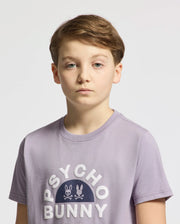 A young person with short brown hair wears a Psycho Bunny KIDS GOLDIE GRAPHIC TEE in lavender, showcasing the "Psycho Bunny" logo and bunny graphic. Made from soft Peruvian Pima cotton, they stand against a plain light background.