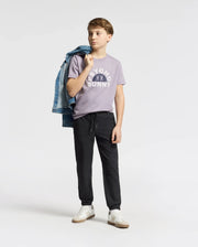 A young person stands against a plain background wearing a purple KIDS GOLDIE GRAPHIC TEE by Psycho Bunny, black pants, and white sneakers. They drape a blue denim jacket over one shoulder and gaze thoughtfully to the side.