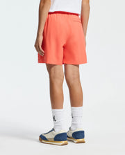 The individual wears Psycho Bunny's KIDS SAMOS HYDROCHROMIC PRINTED SWIM TRUNK - B0W453F200 with white socks featuring black designs and sneakers accented in blue and beige. They stand facing away, highlighting the swim trunk's quick-dry fabric fit against a plain backdrop.
