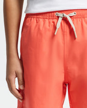 A child is wearing Psycho Bunny's KIDS SAMOS Hydrochromic Printed Swim Trunk - B0W453F200 in coral, featuring quick-dry fabric and an elastic waistband with a light-colored string, paired with a white top. Their right hand rests relaxed by their side.