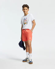 A teen boy, dressed in a casual and sporty outfit, wears a white T-shirt with a colorful graphic and Psycho Bunny's Kids Samos Hydrochromic Printed Swim Trunk in coral, paired with sneakers. He's holding a navy cap against a light background that highlights his clothing.