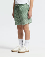 A child stands against a white background wearing Psycho Bunny’s KIDS BENDIGO ALL OVER PRINTED SWIM TRUNK (B0W619F200) in green, paired with a white shirt, white socks featuring a black logo, and white sneakers.