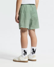 A person stands facing away in Psycho Bunny's KIDS BENDIGO ALL OVER PRINTED SWIM TRUNK (B0W619F200)—green patterned, quick-dry shorts—paired with white socks featuring a black Bunny pattern and white sneakers with brown soles.