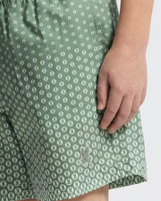 A close-up shows a child wearing Psycho Bunny KIDS BENDIGO ALL OVER PRINTED SWIM TRUNK—green with white bunny and leaf print. Their right hand rests near the pocket, highlighting the quick-dry fabric perfect for active days.