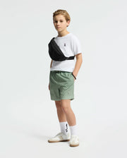 A boy stands against a white background wearing a white t-shirt, Psycho Bunny KIDS BENDIGO ALL OVER PRINTED SWIM TRUNK in green, white sneakers, patterned socks, and a black crossbody bag, looking to the side with his hands in his pockets.
