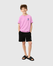 A young boy with curly hair is wearing a light pink t-shirt featuring a small logo on the left chest area, black Psycho Bunny Kids Parker Hydrochromic Swim Trunk (B0W646C200) with a small logo near the hem, and black slides. With his hands in his pockets, he is smiling at the camera against a plain background.
