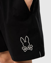 A person wearing the Psycho Bunny KIDS PARKER HYDROCHROMIC SWIM TRUNK (B0W646C200), which features a white skull and crossbones design with a bunny head, is shown with their hand in the pocket. The plain, light-colored background highlights the quick-dry material of the shorts, ideal for any adventure.