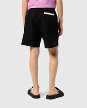 A person is shown from the back, wearing Psycho Bunny's KIDS PARKER HYDROCHROMIC SWIM TRUNK - B0W646C200, featuring black quick-dry material with an elastic waistband and a white strip on the pocket, paired with a pink shirt and black slide sandals against a plain light gray background.
