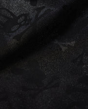Close-up of Psycho Bunny's KIDS PARKER HYDROCHROMIC SWIM TRUNK (B0W646C200) featuring a subtle, intricate pattern of skulls, bones, and abstract shapes that creates a textured, shadowy effect on the black fabric. The quick-dry material boasts a glossy finish with a slightly shimmering surface.